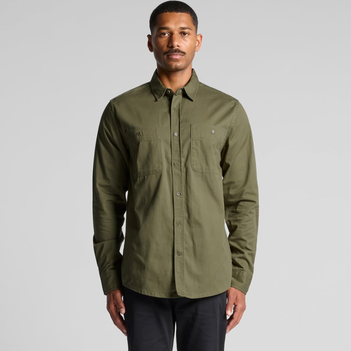 Man standing wearing an olive green button-up shirt and black pants against a plain background.