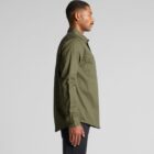Man in olive green button-up shirt and black pants standing in profile against gray background.