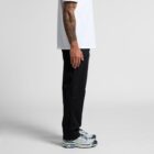Person standing sideways in black pants, white shirt, and sneakers against a plain background.