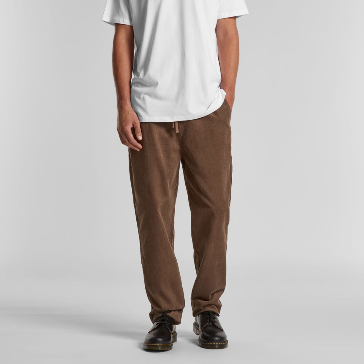 Person wearing a white t-shirt, brown pants, and black shoes standing against a gray background.