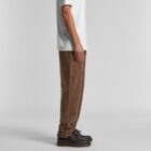 Man in white t-shirt, brown pants, and black shoes standing in profile against plain background.