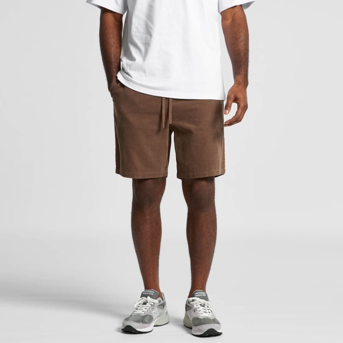 Man wearing a white t-shirt, brown shorts, and gray sneakers stands against a plain background.