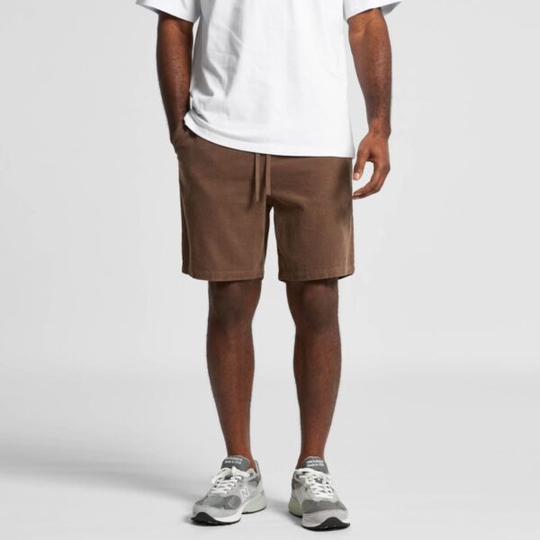Man wearing a white t-shirt, brown shorts, and gray sneakers, standing against a plain background.