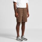 Man wearing a white shirt, brown shorts, and gray sneakers, standing against a plain background.