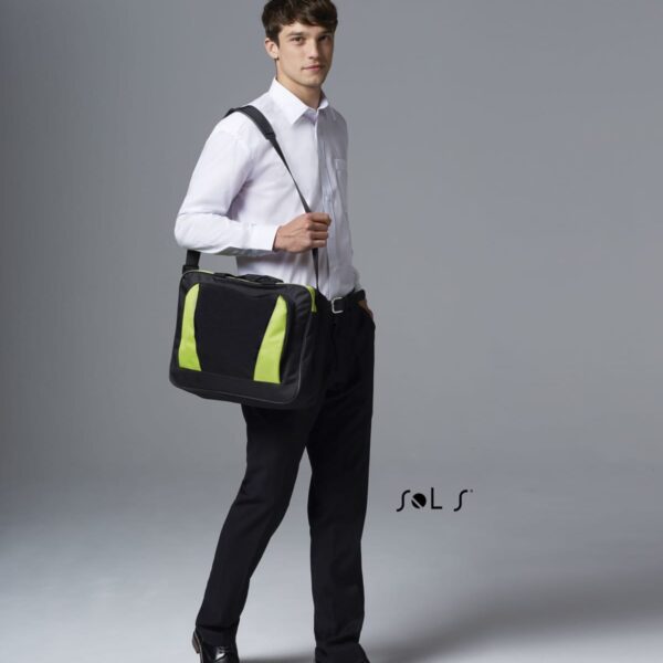 Man in white shirt and black pants holding a black and green shoulder bag, standing indoors.