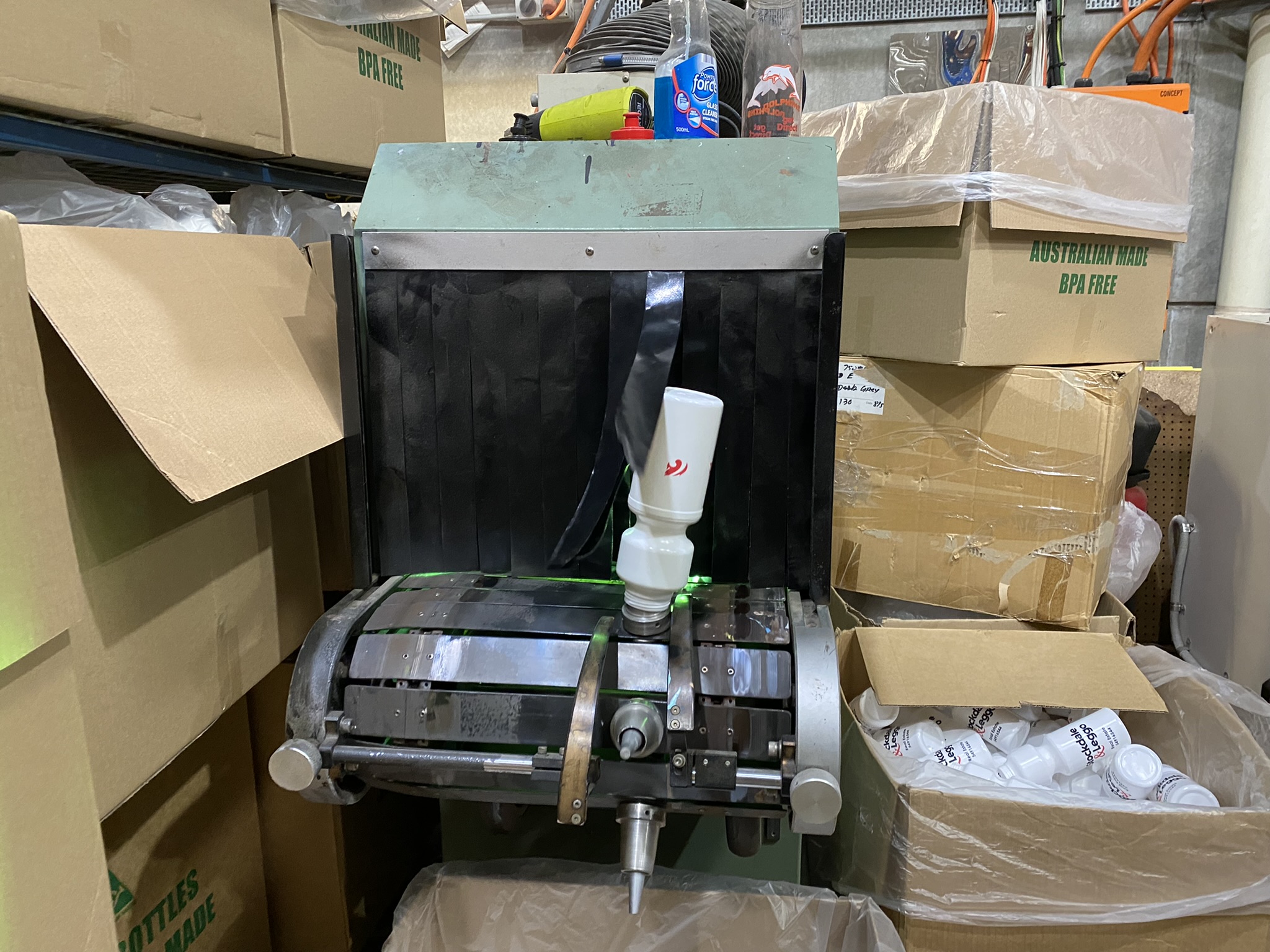 Plastic bottle labeling machine in a warehouse with Custom Bottles and empty boxes nearby.