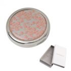 Round silver compact mirror with a pink floral pattern, next to a small white Cacharel box.