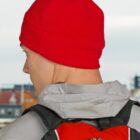 Person wearing a red beanie and gray hoodie with a red and black backpack, seen from behind.