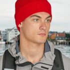 Man in red beanie and gray jacket with backpack, standing outdoors near water and buildings.