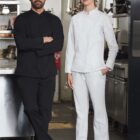 Two chefs in a kitchen, one in black uniform and one in white, standing side by side.