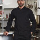 A man in a black chefs uniform stands confidently in a professional kitchen.