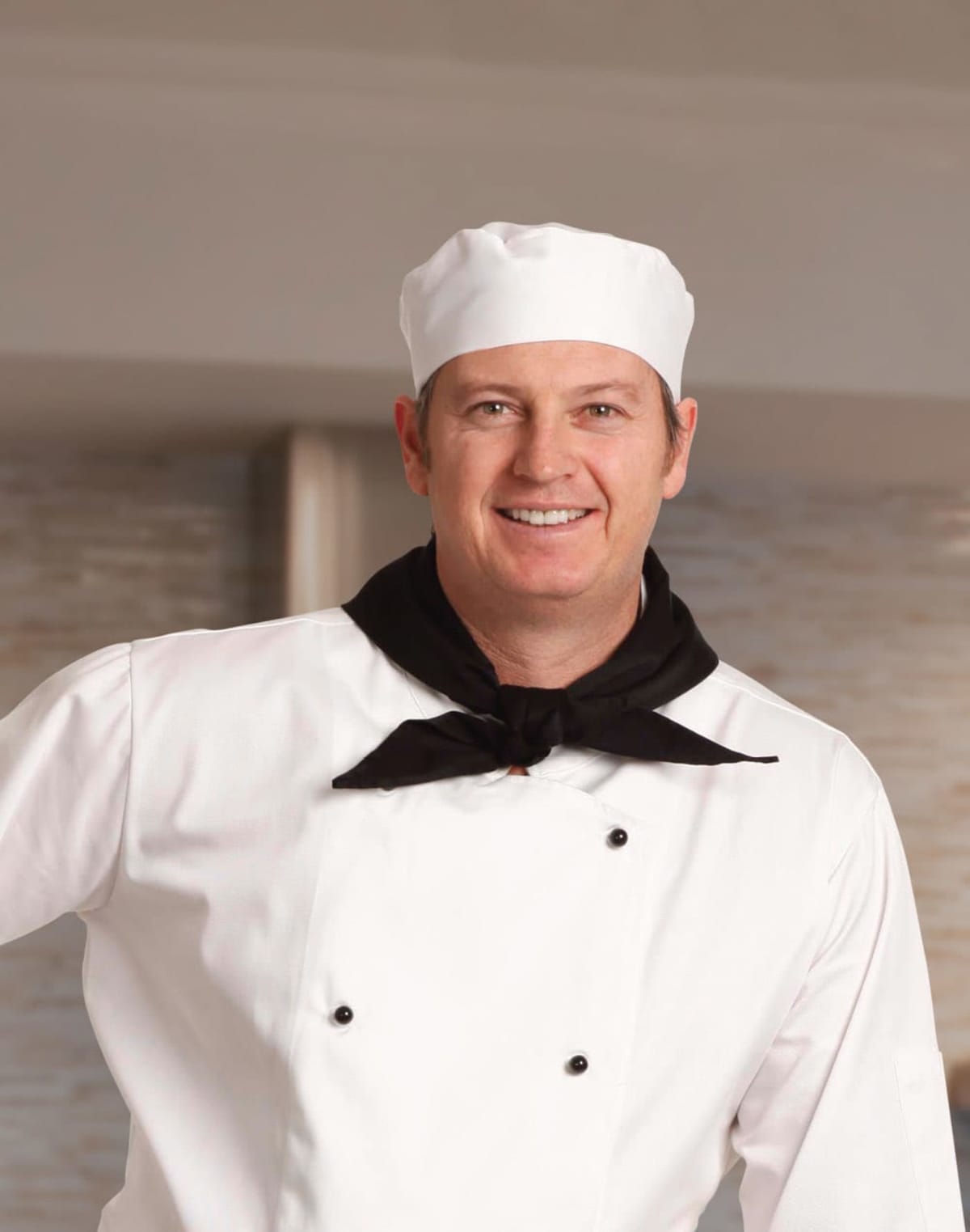 A smiling chef in a white uniform and hat with a black neckerchief.