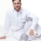 Man in a white hoodie and pants sits on the floor, smiling against a white background.