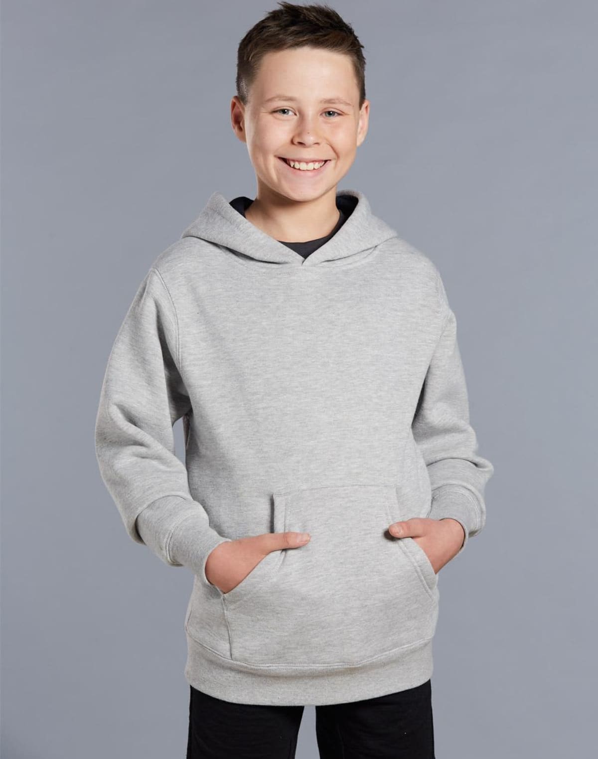 Smiling boy in a light gray hoodie with hands in pockets, standing against a gray background.
