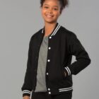Smiling person wearing a black varsity jacket over a gray shirt against a gray background.