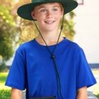 Smiling boy in a blue shirt and green sun hat standing outdoors.