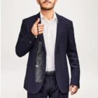 Man in a suit holding a closed umbrella by its strap against a plain background.