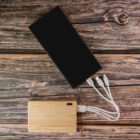 A black smartphone and a wooden power bank connected by three white charging cables on wood surface.