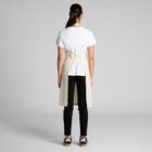 Person in a white shirt, beige apron, and black pants standing with back facing the camera.