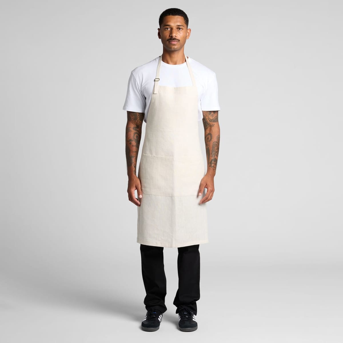 Man wearing a beige apron over a white shirt and black pants, standing against a gray background.