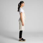 Woman in apron, white shirt, black pants, and sneakers standing in profile against gray background.