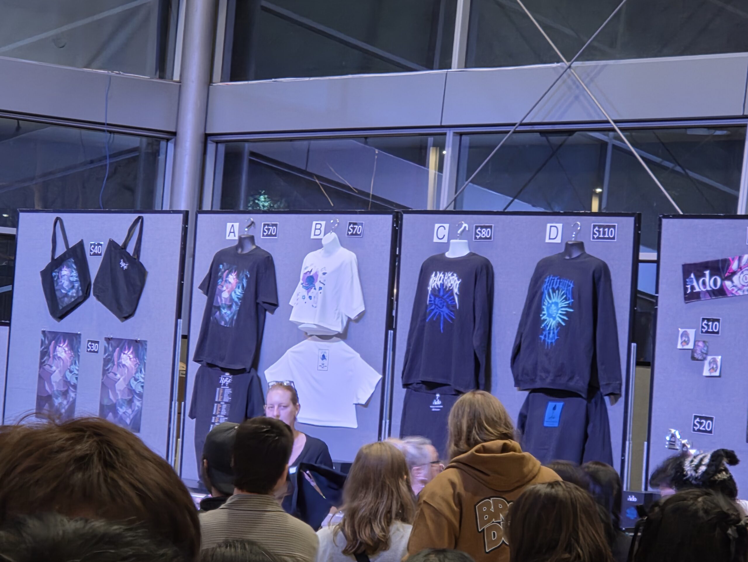 People shop at a booth displaying concert merchandise like shirts, bags, and posters.