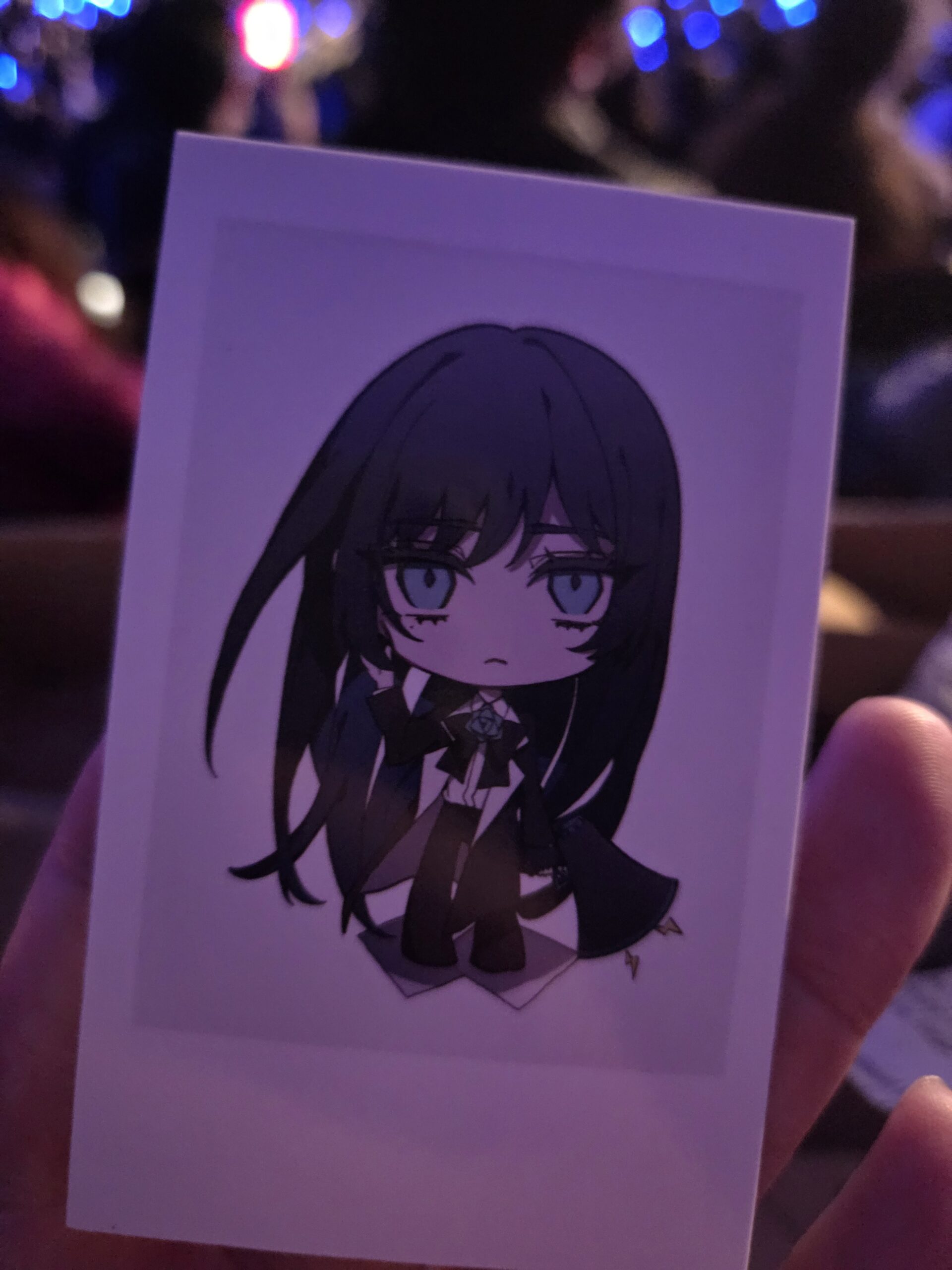 A hand holds concert merchandise—a chibi anime-style drawing of a sad girl with long dark hair.
