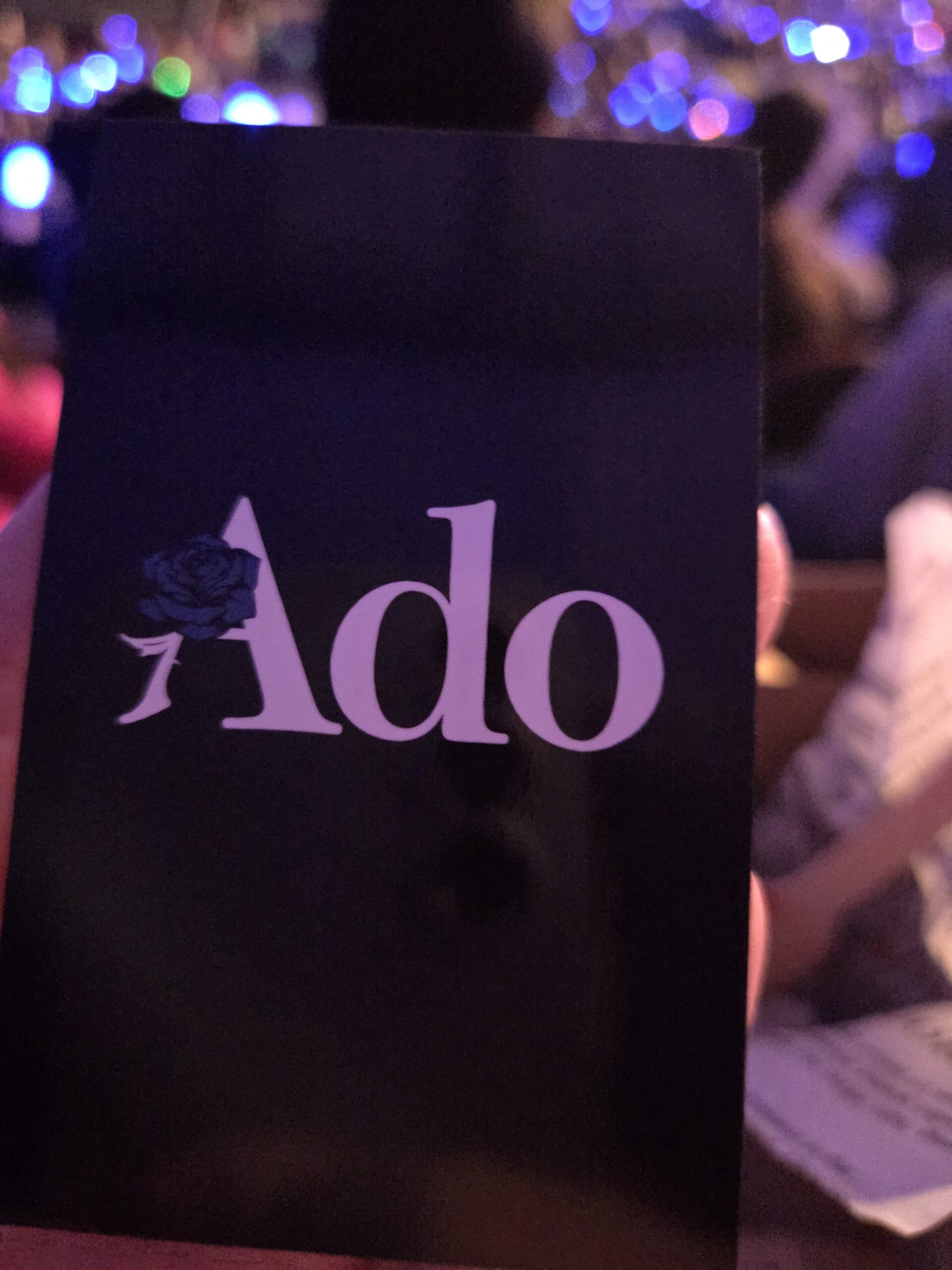 A hand holds concert merchandise: a card with Ado and a blue rose amid blurred lights.