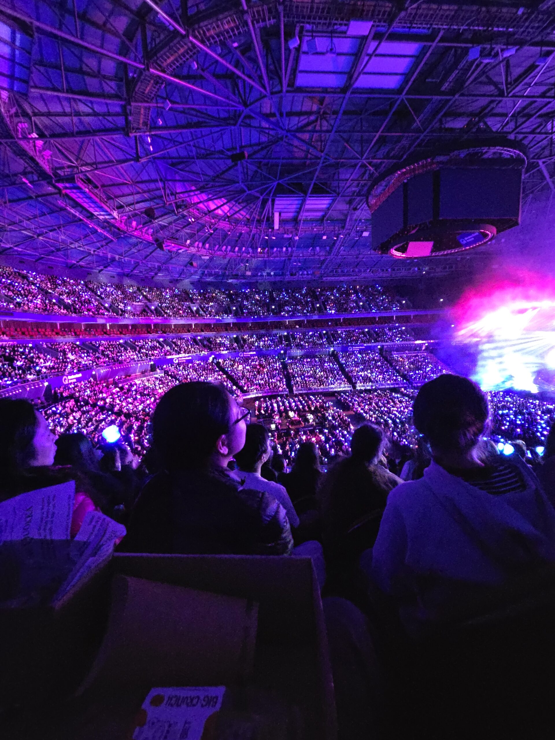 Large indoor concert crowd with glowing lights and fans showing off concert merchandise.