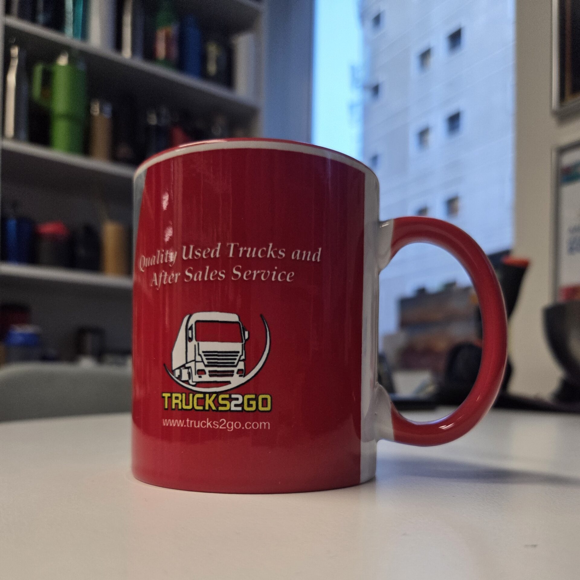 Red Trucks2Go ceramic mug on a white desk with office shelves and a window in the background.