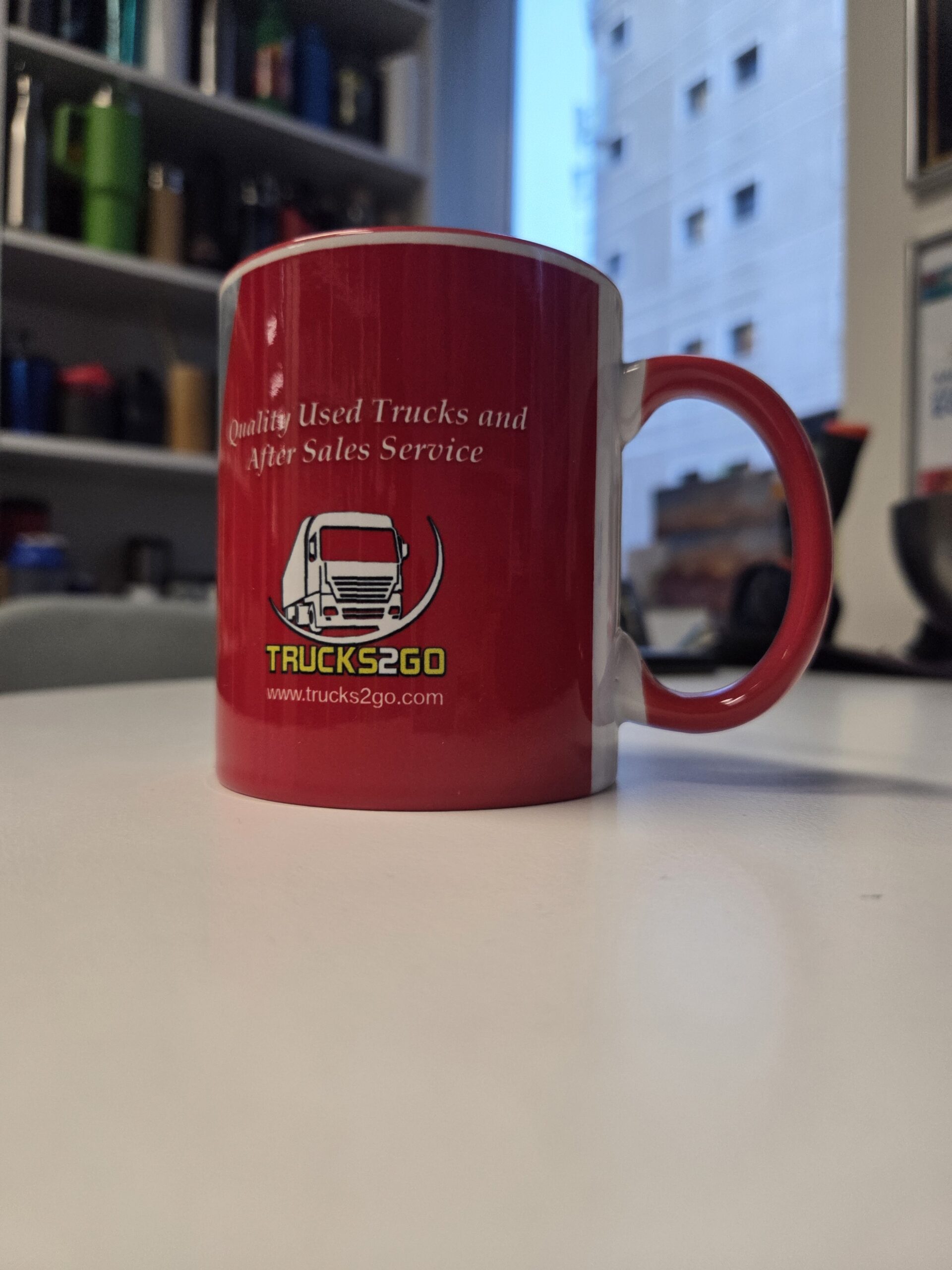Red Trucks2Go ceramic mug on a white desk with office shelves and a window in the background.