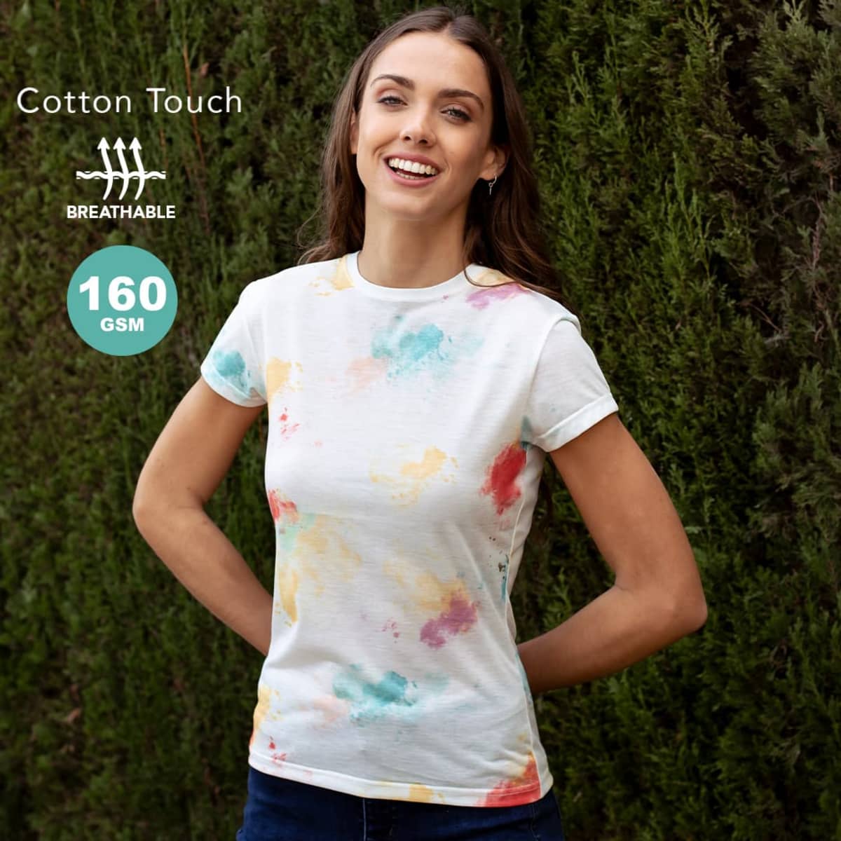 Woman wearing a colorful cotton t-shirt stands outside in front of green bushes, smiling.