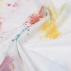 White fabric with colorful paint stains in yellow, red, green, and purple, loosely wrinkled.
