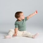 Toddler in green shirt sits on floor, reaching up with one hand, looking at fingers.