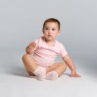 Baby in a pink onesie and socks sitting on the floor against a plain gray background.