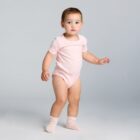 Toddler in a light pink onesie and socks standing against a plain gray background.