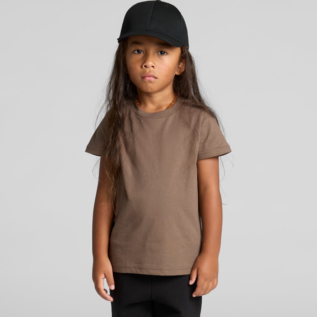 Child with long hair wearing a brown t-shirt and black cap stands against a plain background.