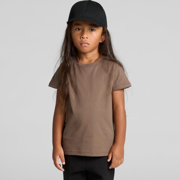 Child with long hair, brown t-shirt, black cap, and black pants standing against gray background.