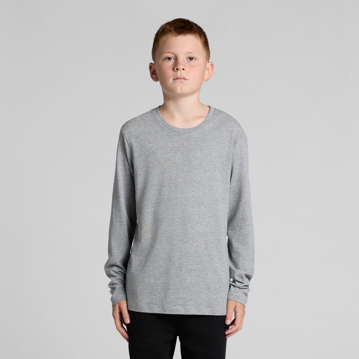 A boy in a gray long-sleeve shirt and black pants stands against a plain background.
