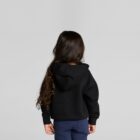 A child with long hair in a black hoodie stands facing away against a plain background.