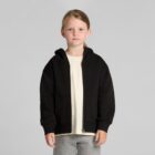 A young girl in a black hoodie, cream shirt, and gray pants stands against a plain background.