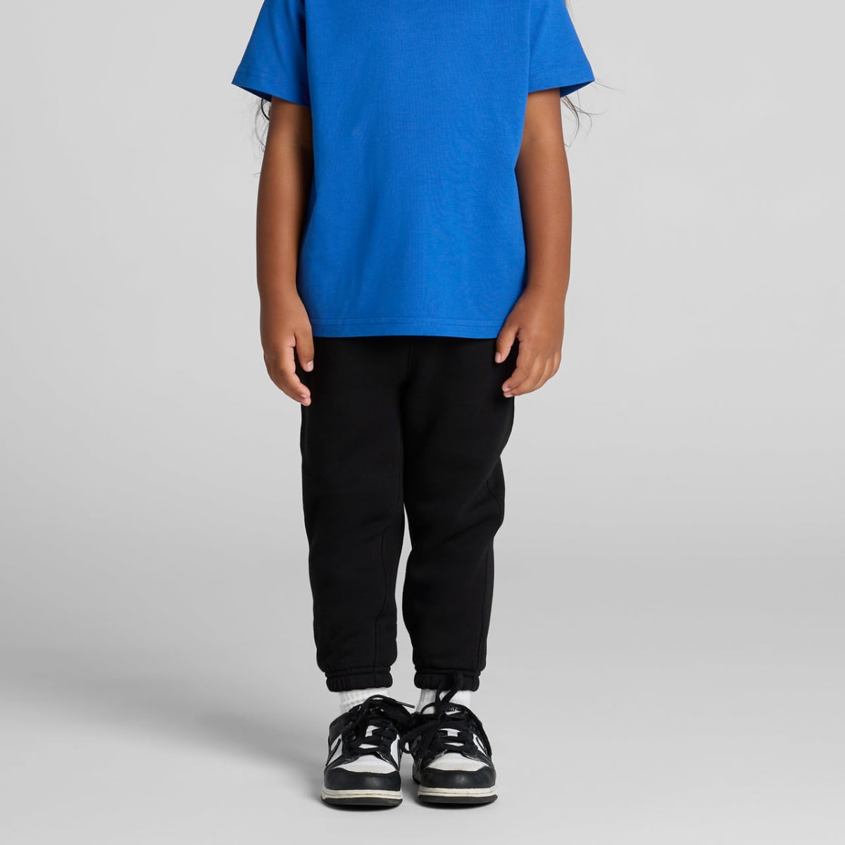 Child wearing a blue T-shirt, black pants, and black sneakers standing against a gray background.