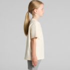 Young girl with a ponytail in a cream t-shirt and gray shorts standing in profile.