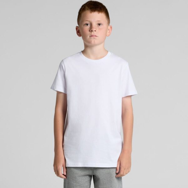 A boy in a plain white t-shirt and gray pants stands facing forward against a gray background.