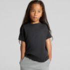 A child in a black t-shirt and gray pants stands with hands in pockets against a plain background.