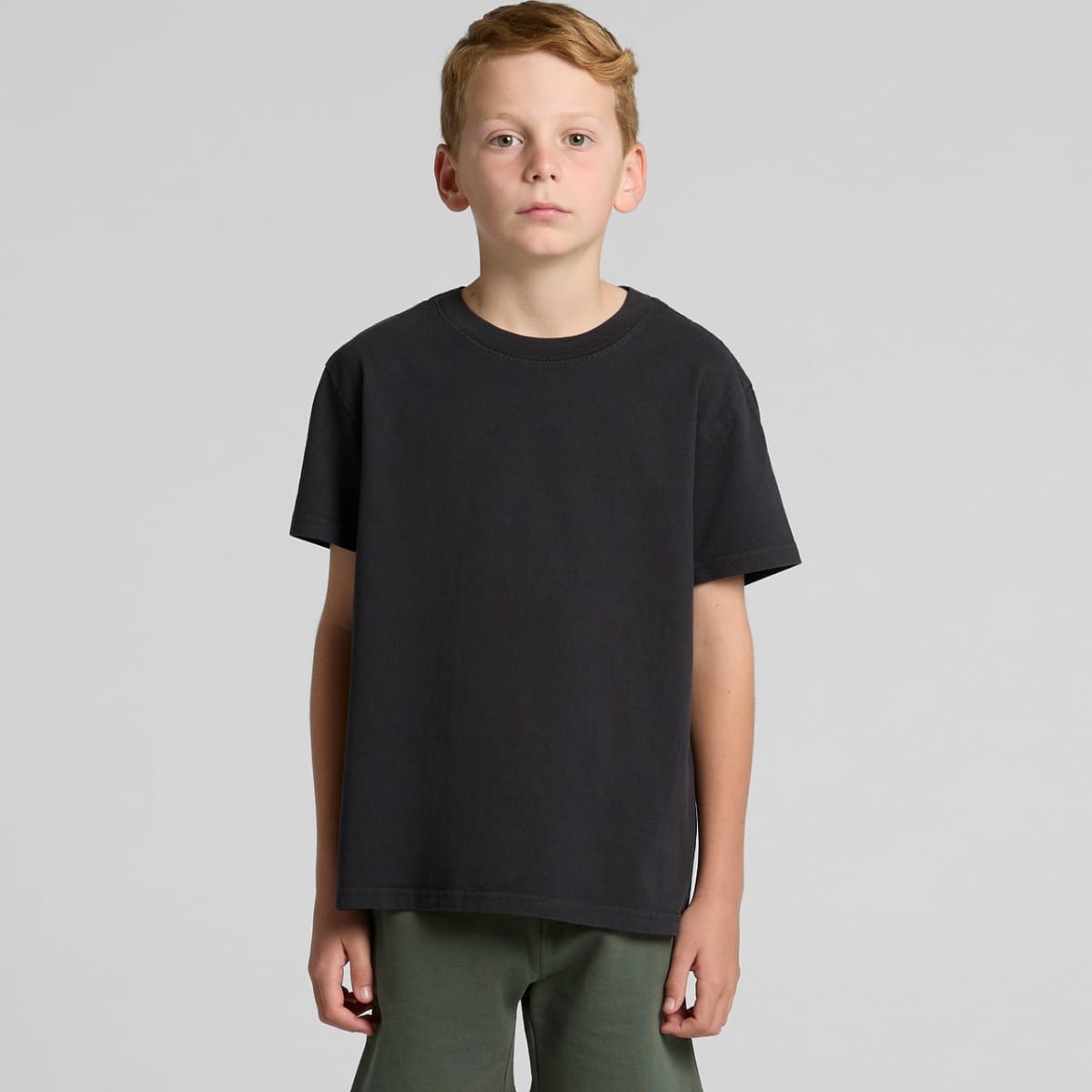 Young boy with short hair wearing a black t-shirt and green pants, standing against gray background.