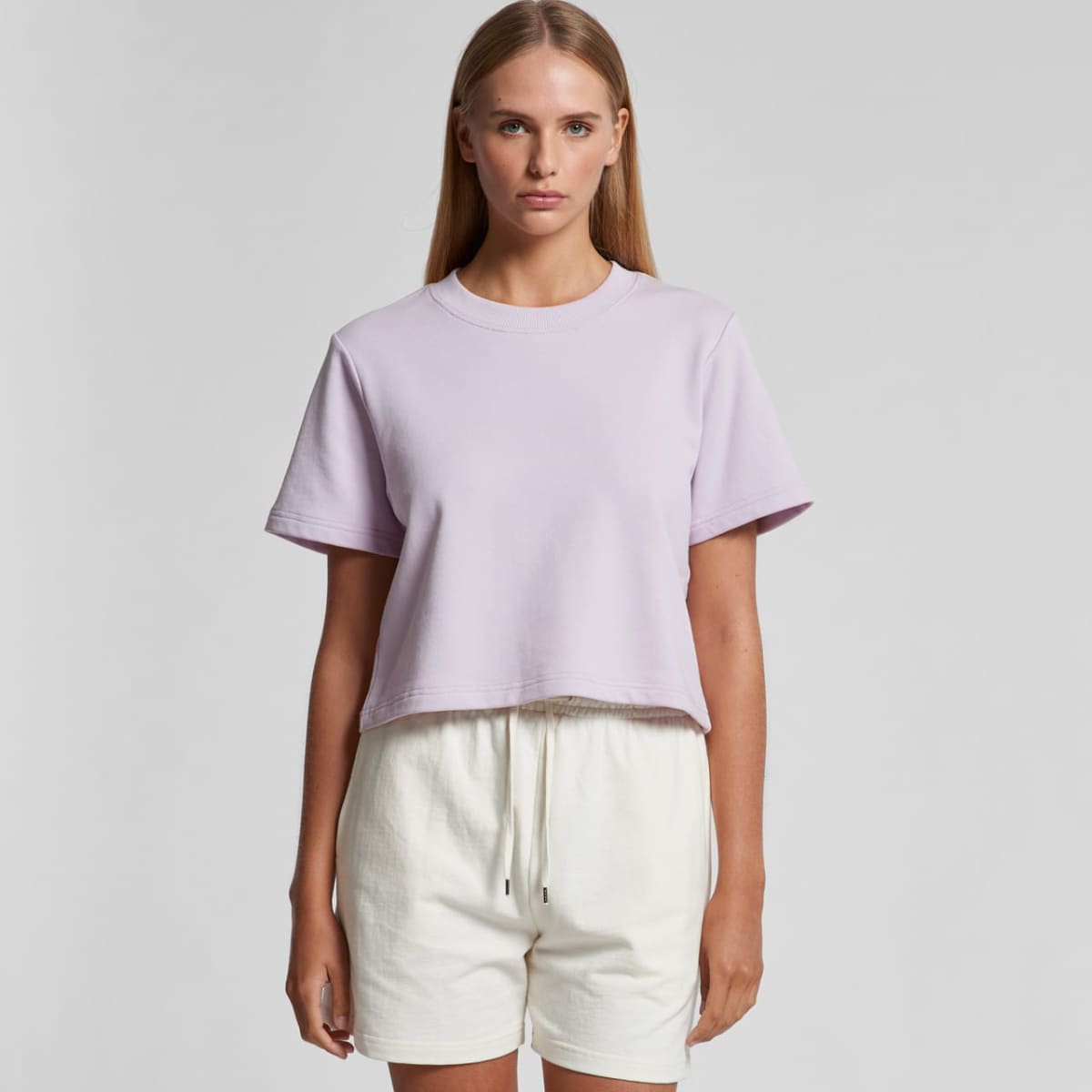 Woman in a light purple t-shirt and white shorts standing against a plain background.