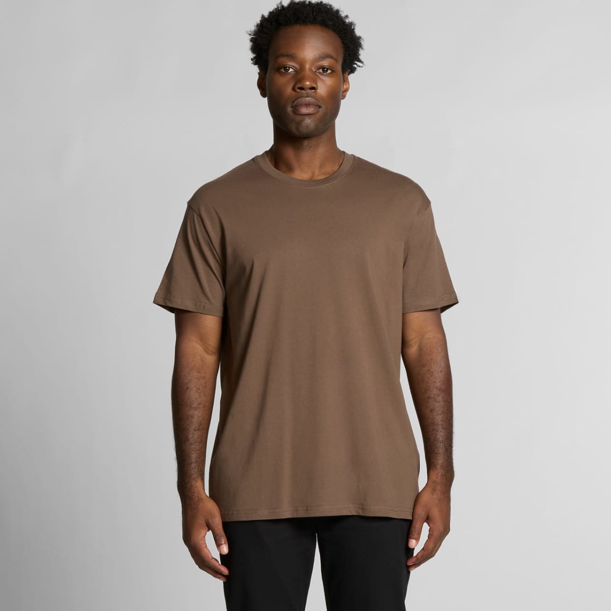Man wearing a plain brown t-shirt and black pants standing against a light gray background.