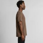 Man standing in profile, wearing a brown t-shirt and black pants against a plain background.