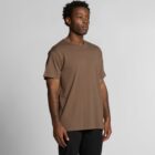 Man wearing a brown t-shirt and black pants standing against a plain gray background.